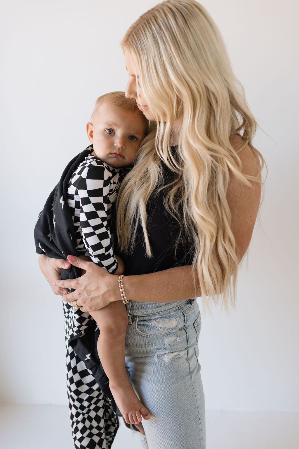 A woman with long blonde hair holds a small child wrapped in a forever french baby Muslin Quilt, featuring Midnight Lightning Bolt and Black + White Wavy Checkerboard patterns. The child gazes towards the camera as the woman, wearing a black top and light blue jeans, looks down at them against a stark white background.