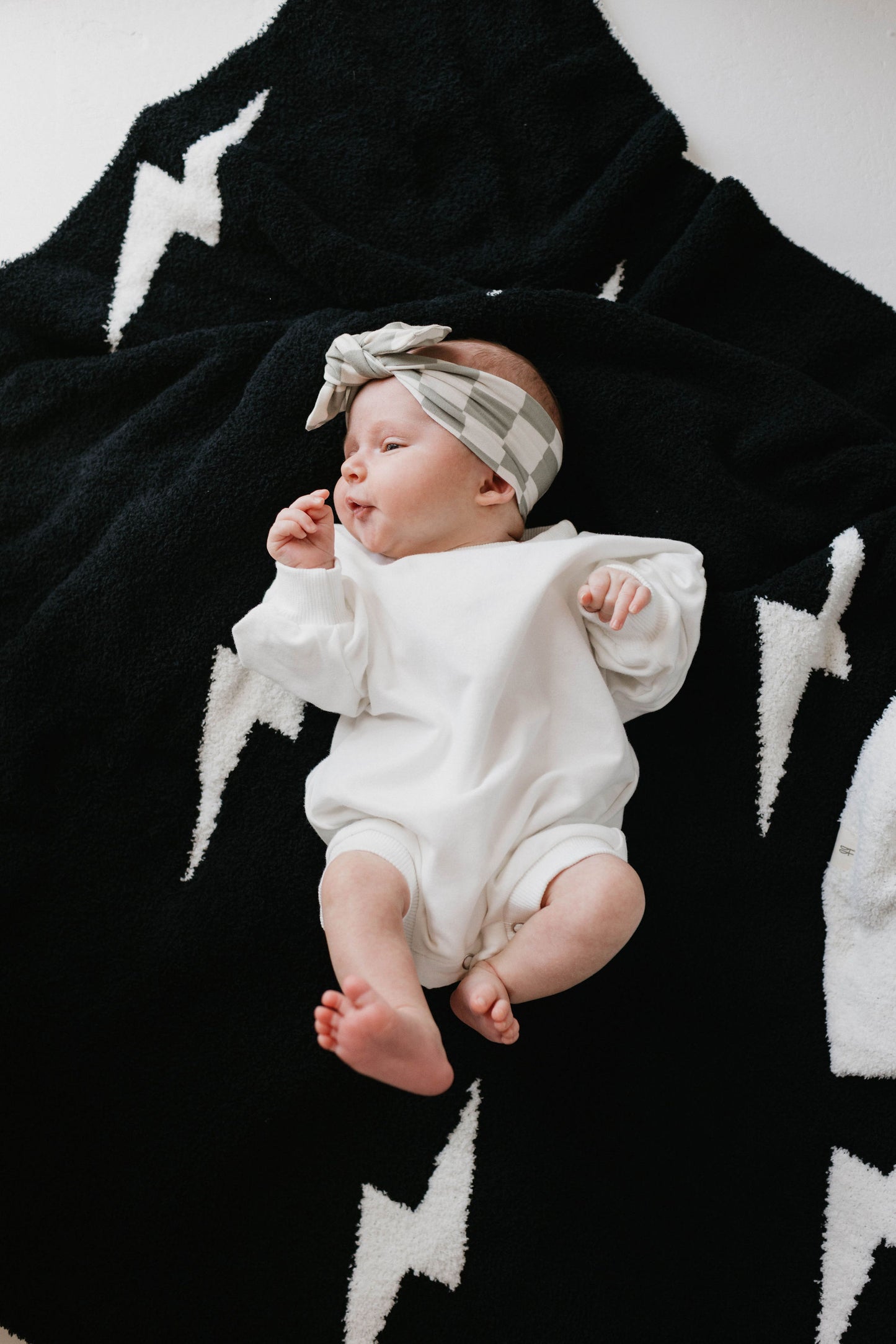 A baby wearing the Sweatshirt Romper | Dove from forever french baby, paired with a checkered headband, lies on a black blanket adorned with white lightning bolt patterns. The baby is positioned slightly to the side and appears to be looking towards the right.