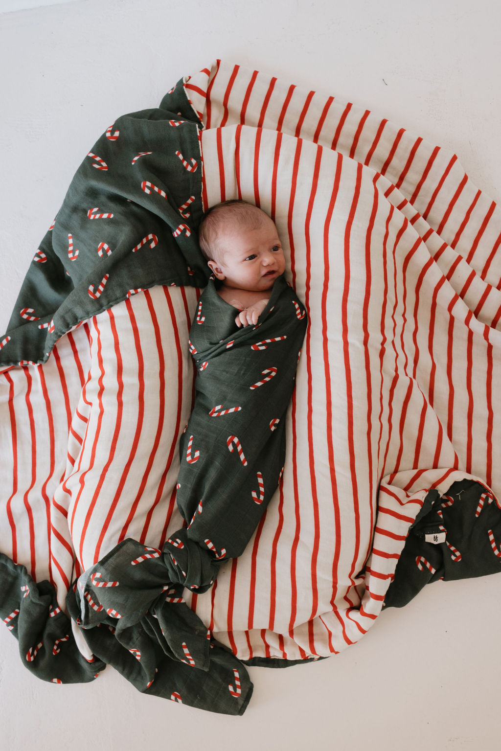 A newborn baby is swaddled in the forever french baby Muslin Swaddle from the Candy Cane Lane collection, resting on a larger red and white striped blanket on a white surface.