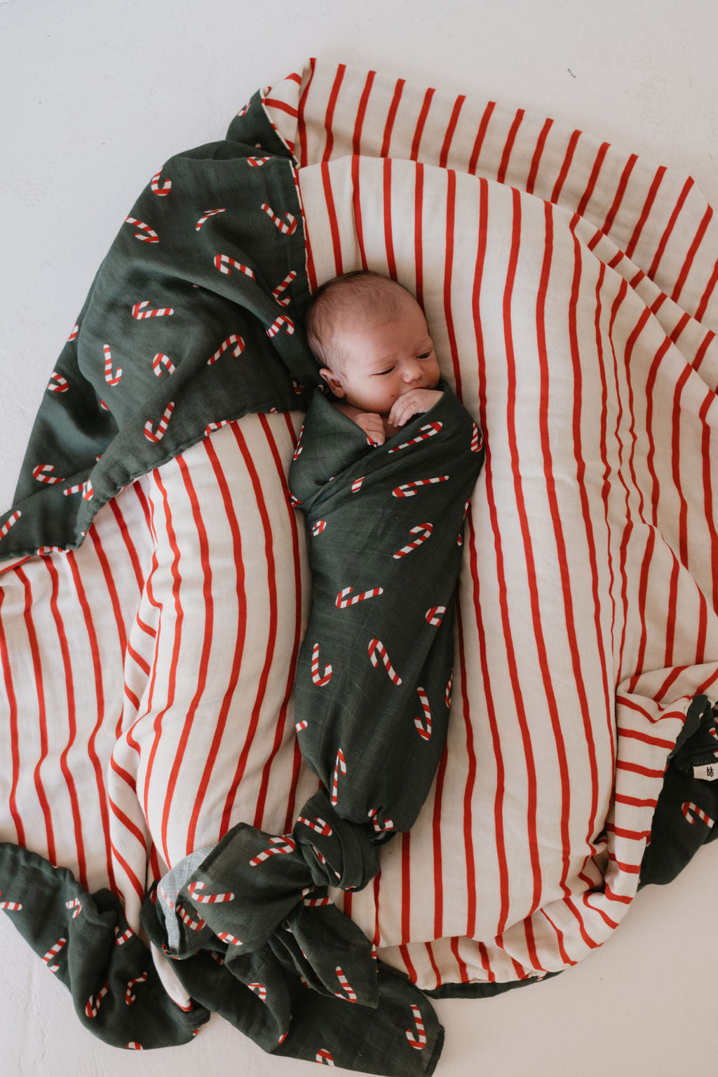 A baby is snugly wrapped in the "Muslin Swaddle | Candy Cane Lane" by forever french baby, featuring festive candy cane patterns on a green background, while lying on a larger red and white striped blanket. The product offers a cozy and peaceful feel.