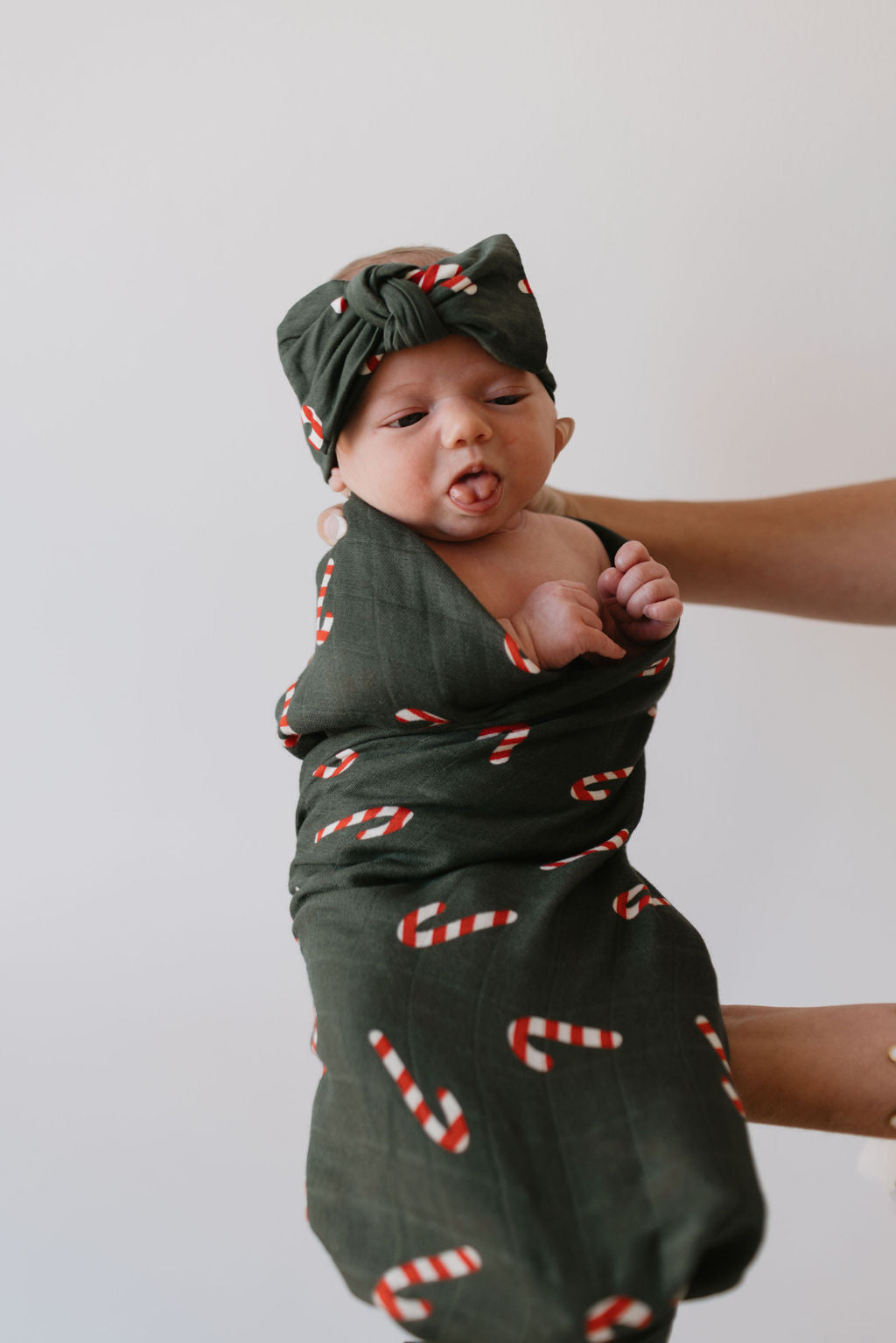 Cradled against a simple white background, a baby cozily wrapped in the 'Candy Cane Lane' muslin swaddle by forever french baby, complete with a matching headband, showcases the charm of dark green cotton adorned with festive candy cane patterns.