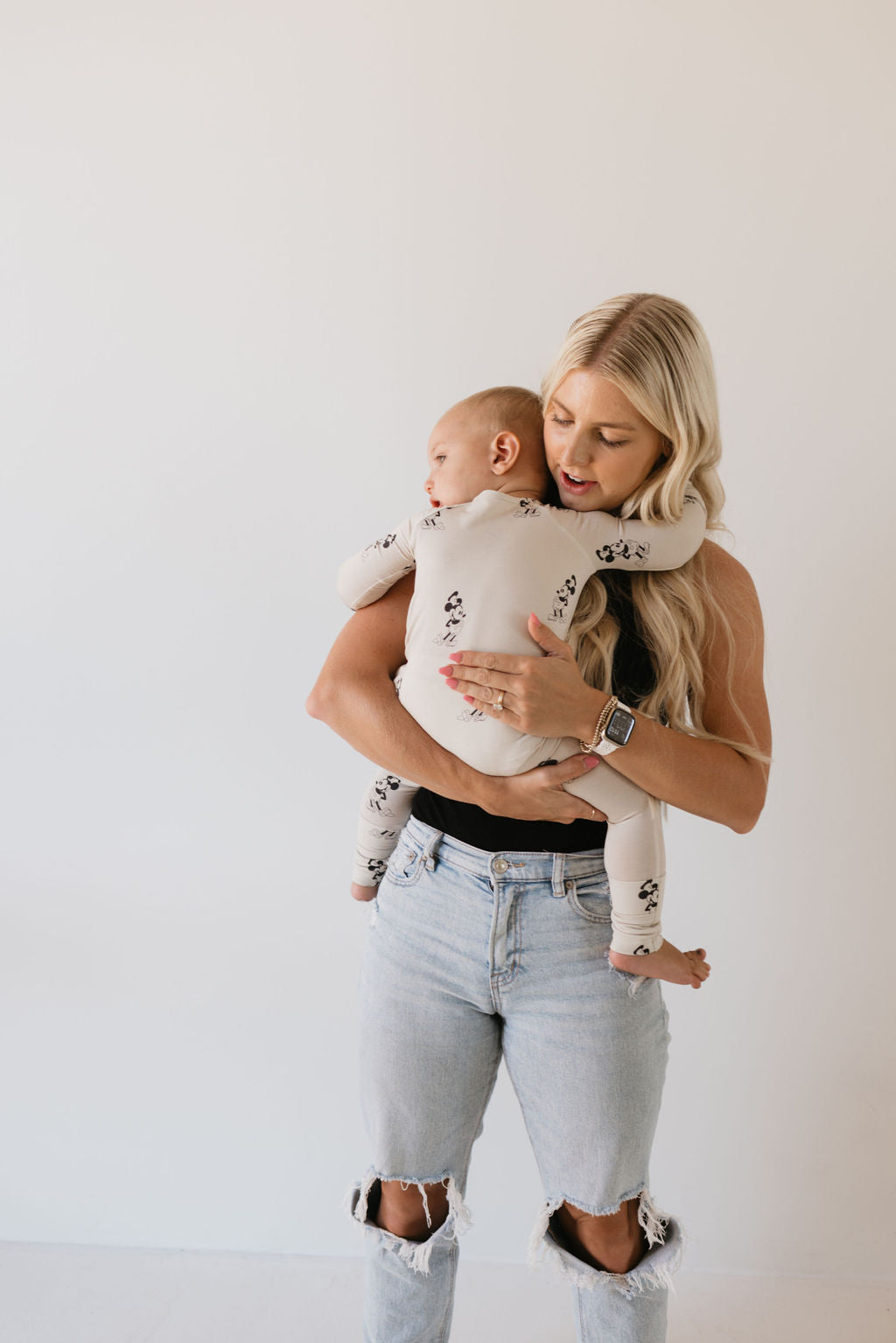 A woman with long blonde hair cradles a baby in bamboo pajamas by Forever French Baby, showcasing the "All Aboard!" design. She is dressed in a sleeveless black top and ripped light blue jeans, standing against a plain white background.