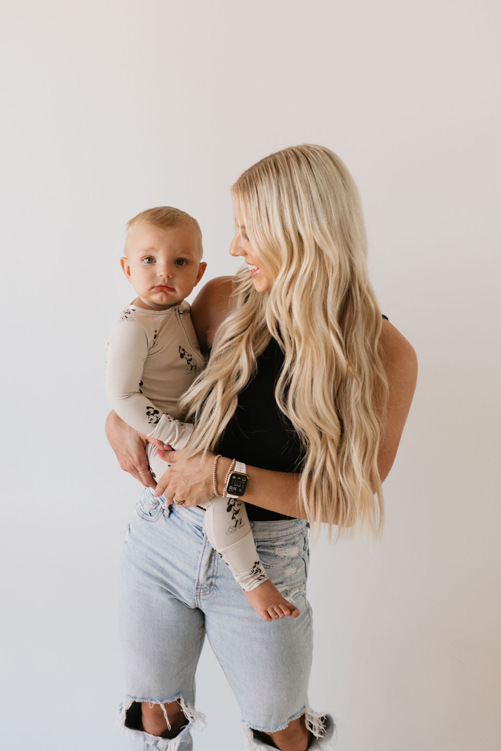 A woman with long blonde hair, dressed in a black top and ripped jeans, gently holds her baby, who is wearing the "Bamboo Zip Pajamas | All Aboard!" from forever french baby. They stand against a plain white background, exchanging soft smiles as they look at each other.