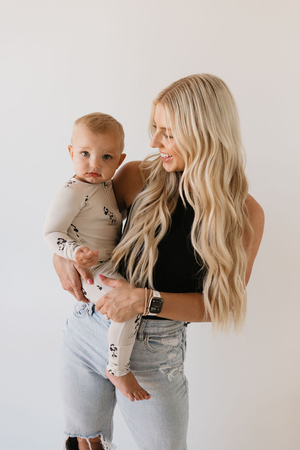 A woman with long blonde hair, dressed in a black sleeveless top and ripped jeans, holds a cheerful baby wearing the forever french baby Bamboo Zip Pajamas in the 'All Aboard!' design. The baby, sporting light brown hair, smiles against a plain white background, highlighting the cozy and breathable sleepwear.