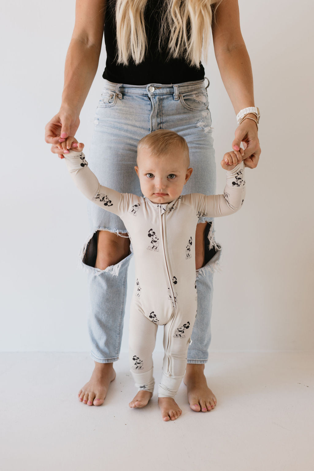 A toddler in a panda-print onesie from the "Bamboo Zip Pajamas | All Aboard!" collection by forever french baby is held up by an adult. The adult is dressed in ripped jeans and a black top against a plain white background.