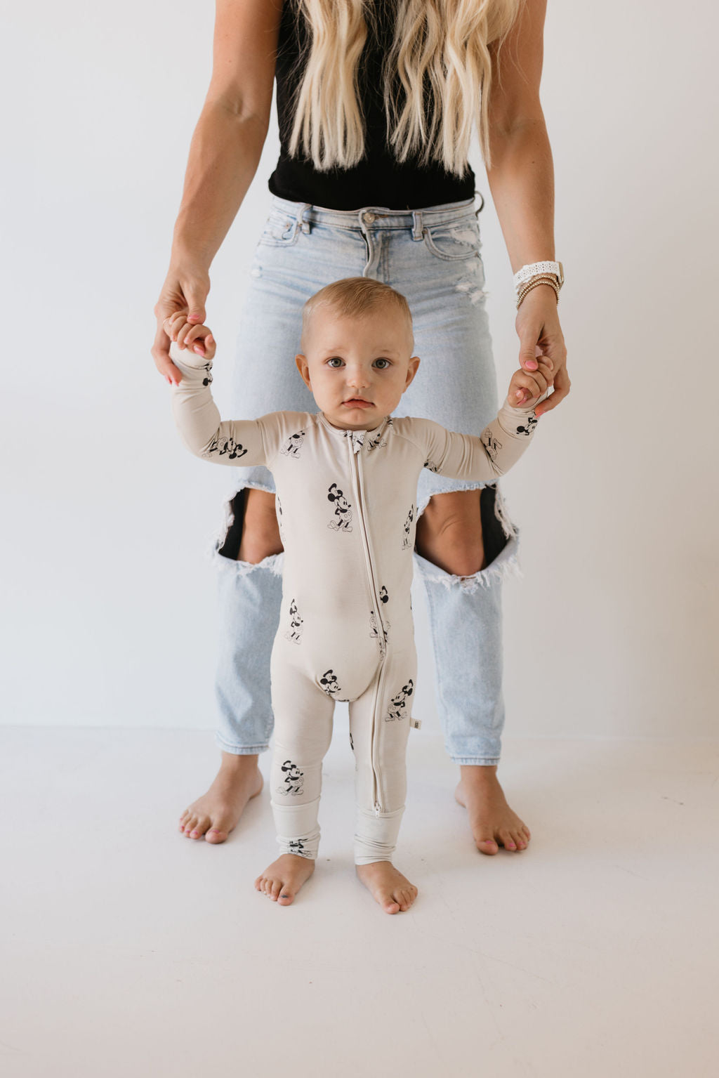 A baby in the "All Aboard!" bamboo zip pajamas by forever french baby, which is cream-colored with small black prints, stands and holds hands with an adult. This hypo-allergenic outfit provides comfort for sensitive skin. The adult, visible from the waist down in a black top and ripped jeans, is set against a white background.