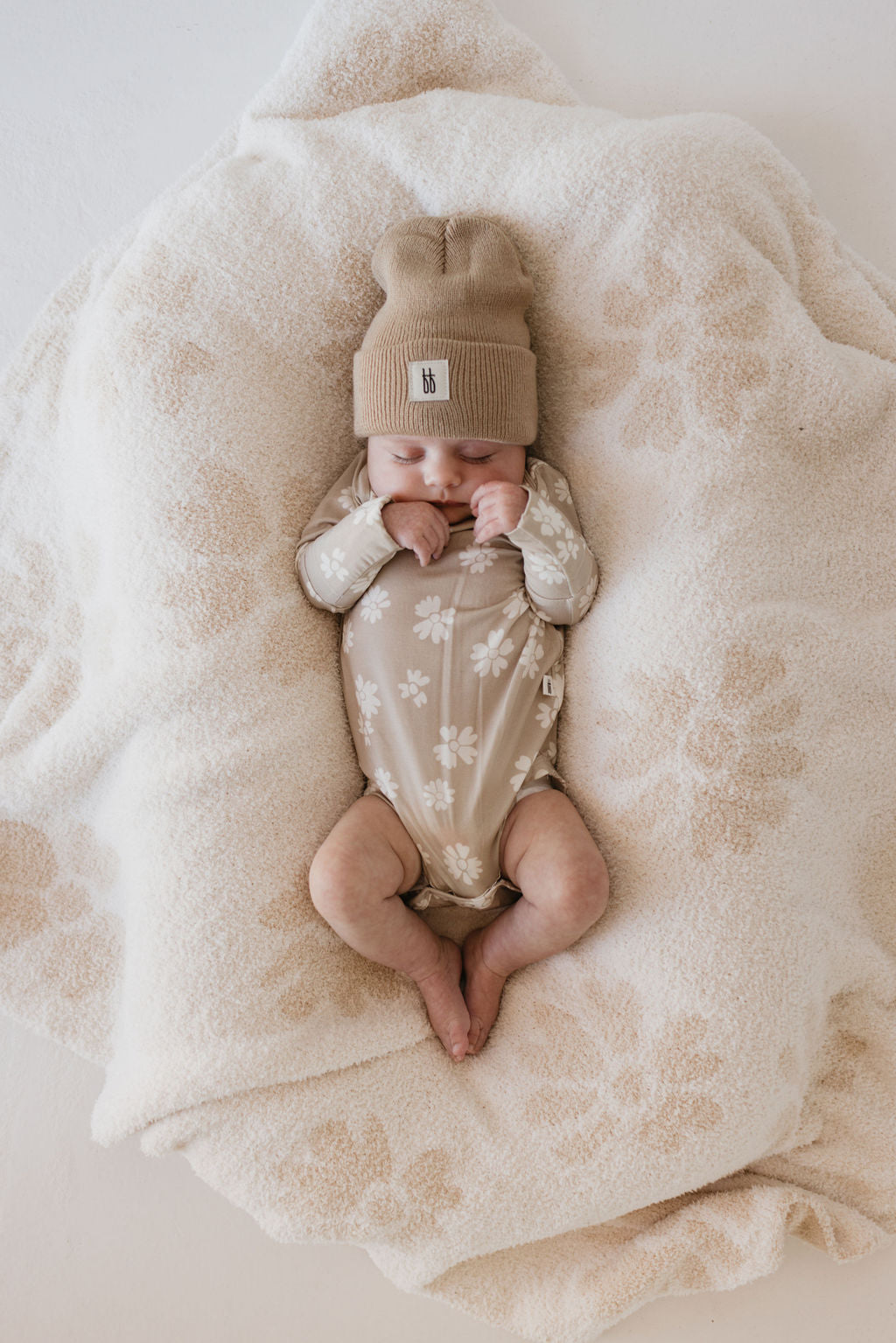 A baby peacefully sleeps on a soft, patterned blanket, wearing forever french baby's Long Sleeve Bamboo Snapsuit in Lazy Daisy and a matching beanie. The bamboo fabric adds to the cozy, serene scene.