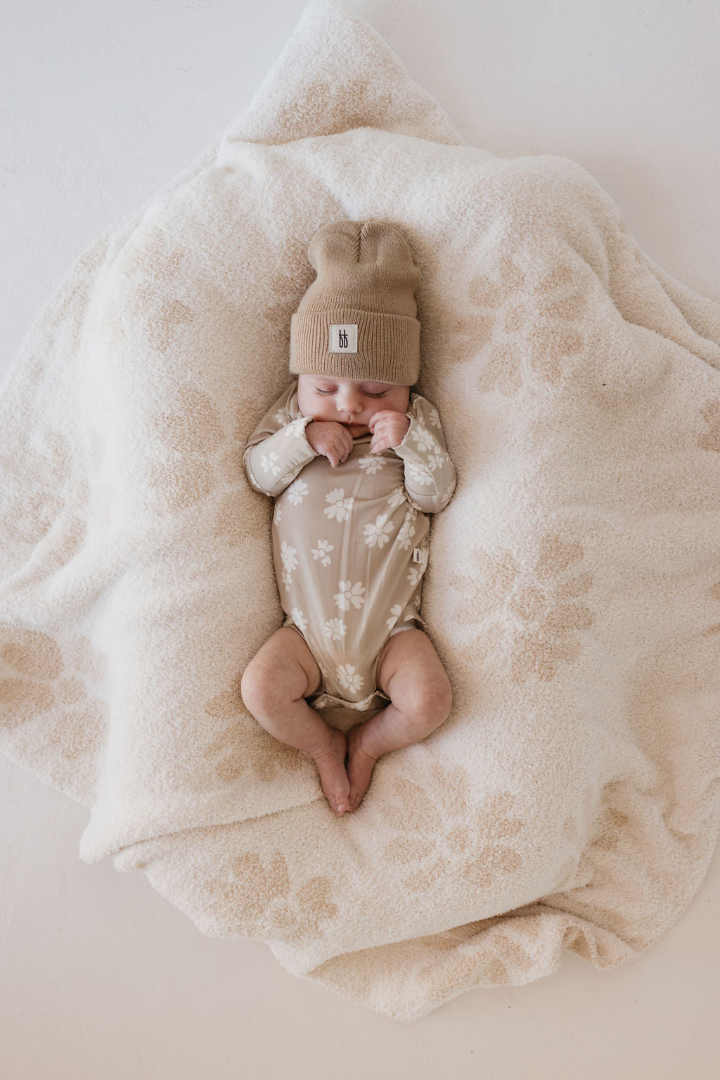 A baby lies on a cozy, patterned blanket of breathable bamboo fabric, wearing forever french baby's Lazy Daisy Long Sleeve Bamboo Snapsuit—beige with white floral designs—and a matching knit hat in a peaceful setting.