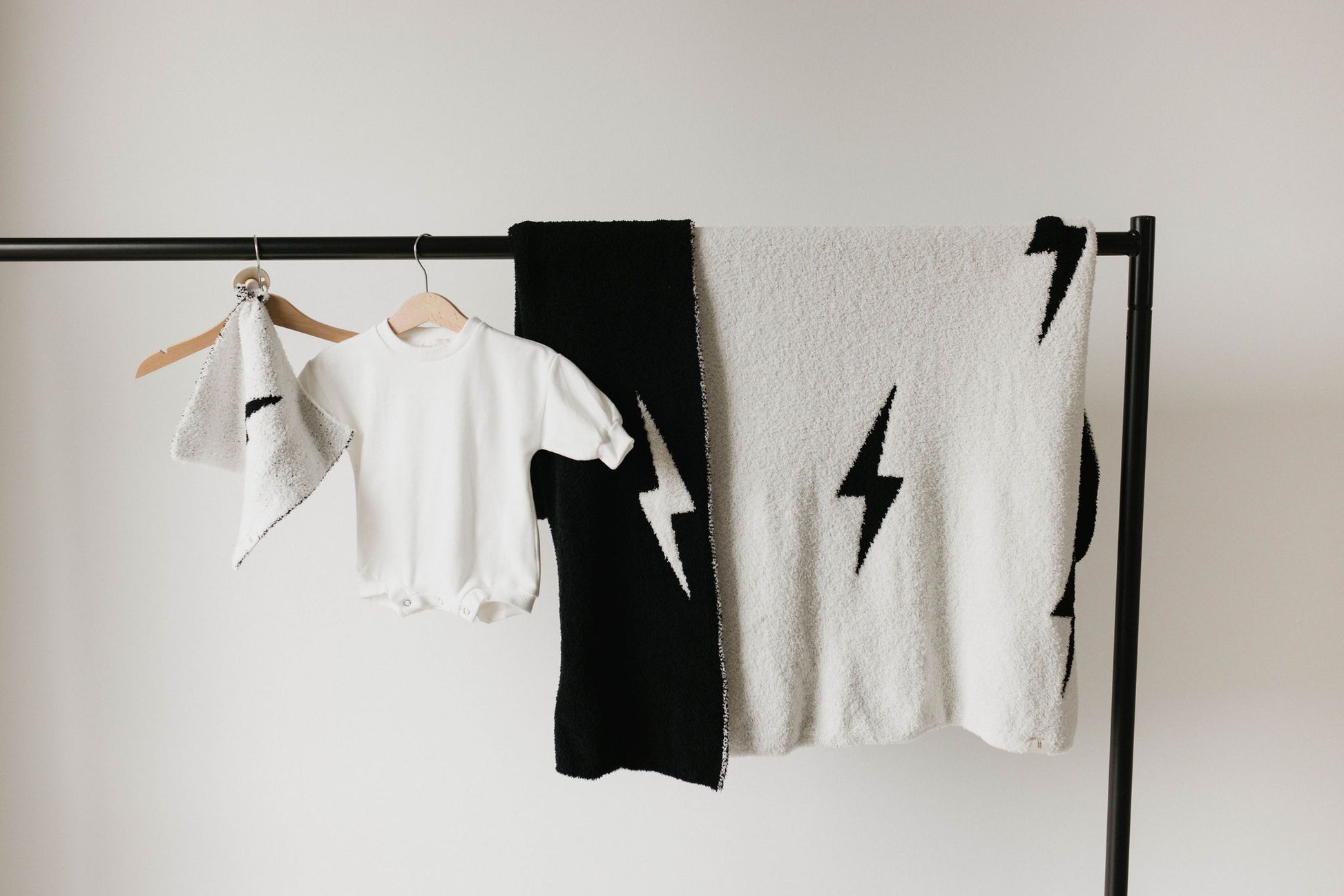 A black metal clothing rack displays a white baby onesie, a knotted small towel in white with black trim, and the forever french baby Plush Blanket featuring a striking black and white lightning bolt pattern. The blanket is large and Oeko-Tex certified.