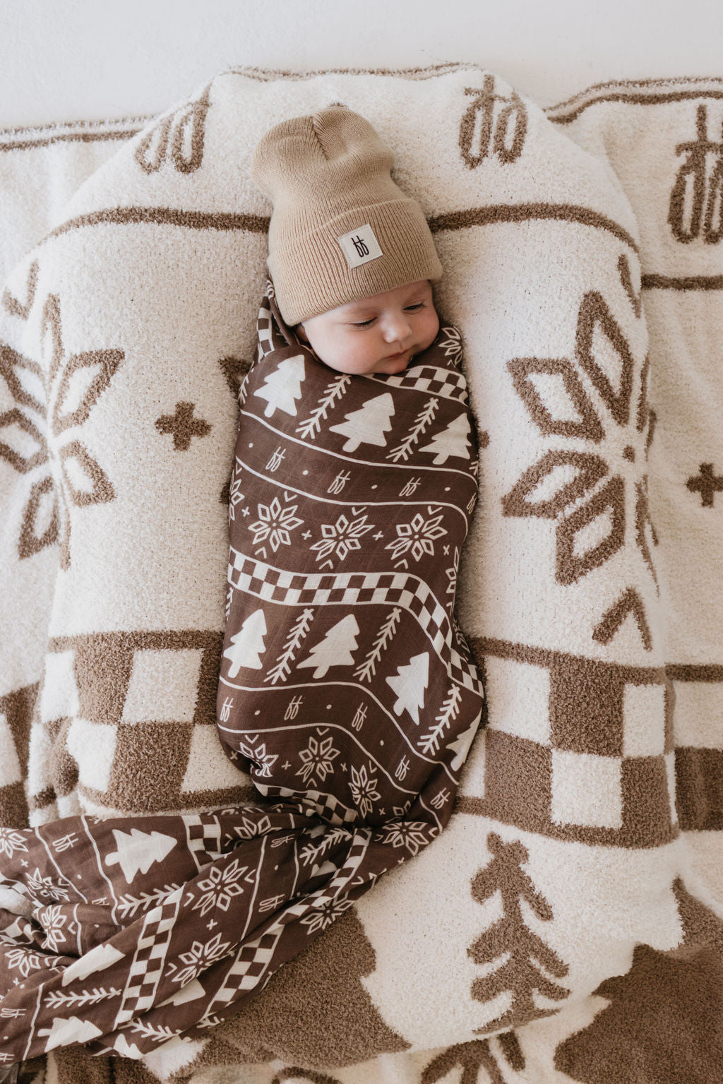 A baby is snuggled in the ultra-soft "Muslin Swaddle | Forever Fair Isle" from forever french baby, showcasing a brown and white pattern with tree and snowflake designs. The cozy muslin fabric perfectly pairs with a beige beanie as the baby rests on a matching fluffy blanket, creating a warm and serene scene.