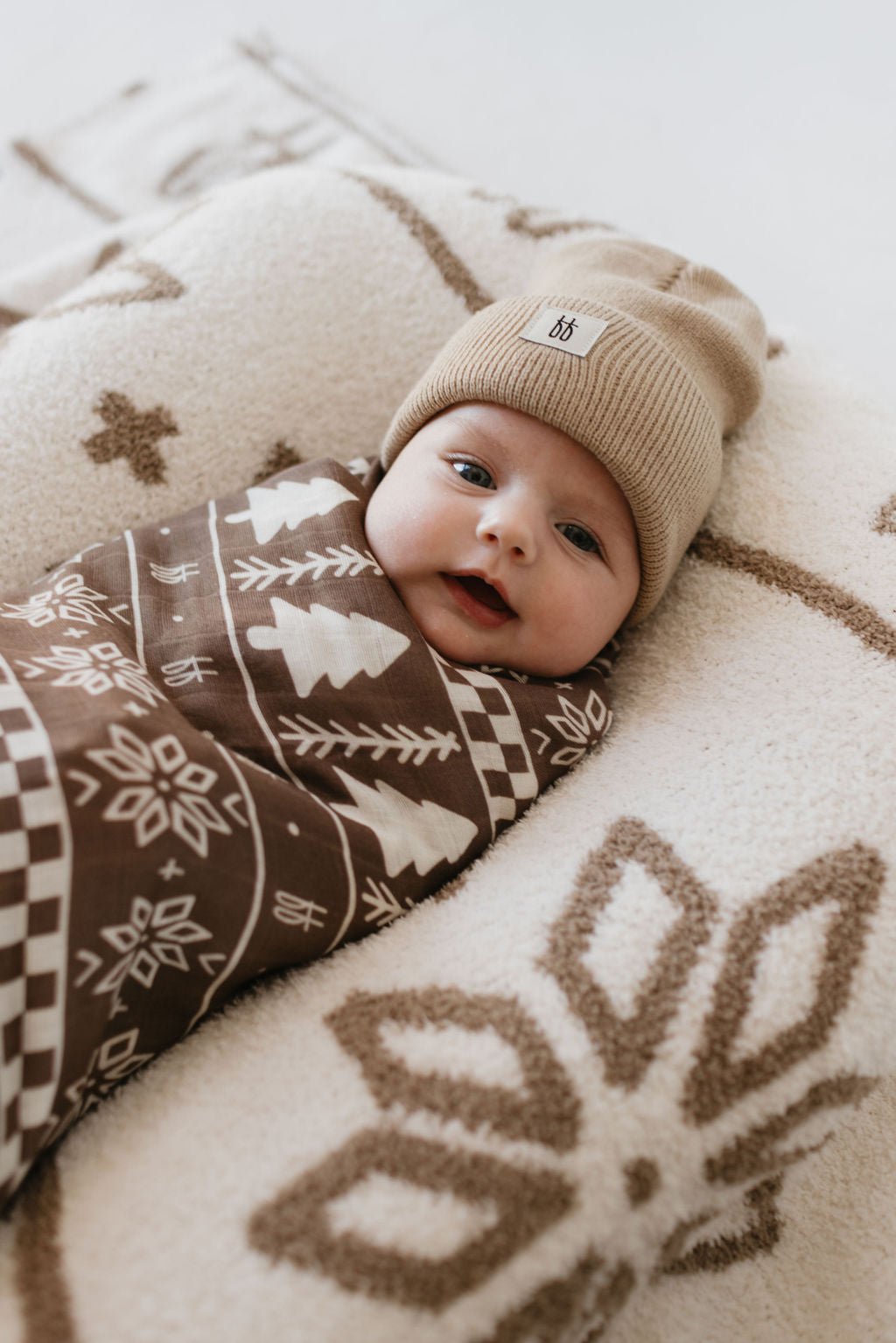 A baby is lying on a fuzzy patterned blanket, wrapped in the ultra-soft "Muslin Swaddle | Forever Fair Isle" by forever french baby, wearing a beige beanie. The background and swaddle feature geometric and tree motifs.