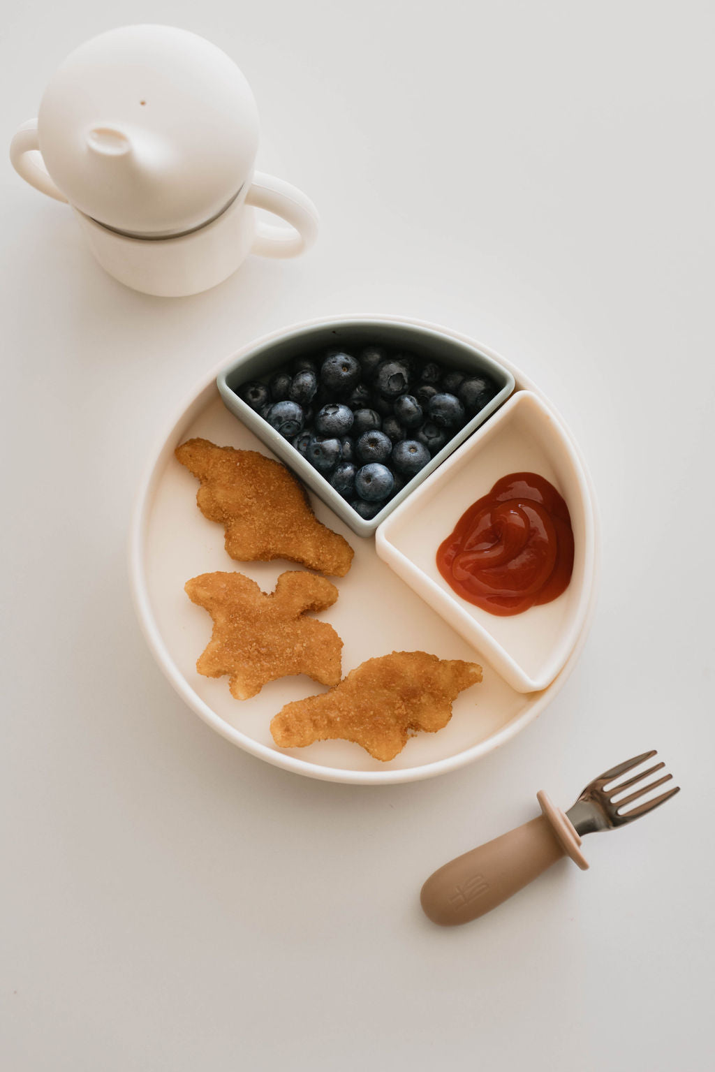 A forever french baby Silicone Plate | Removable Dividers holds compartments of dinosaur-shaped chicken nuggets, fresh blueberries, and a serving of ketchup. A toddler fork with a beige handle is placed nearby, along with a white snack container with a lid. The background is a plain white surface, perfect for mealtime.