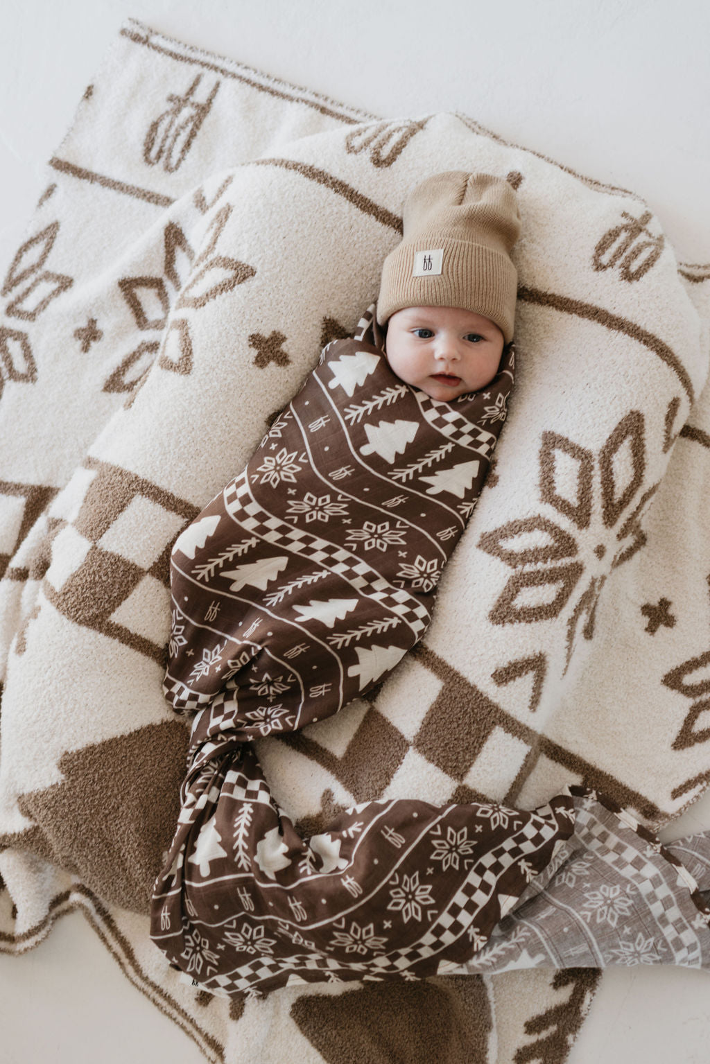 A baby wrapped in the ultra-soft "Muslin Swaddle | Forever Fair Isle" by forever french baby lies on a brown and white patterned mat, both crafted from cozy muslin material. The baby, wearing a beige knit hat, has a peaceful expression. The setting is warm and inviting, emphasizing the intricate patterns and gentle texture of muslin cotton.