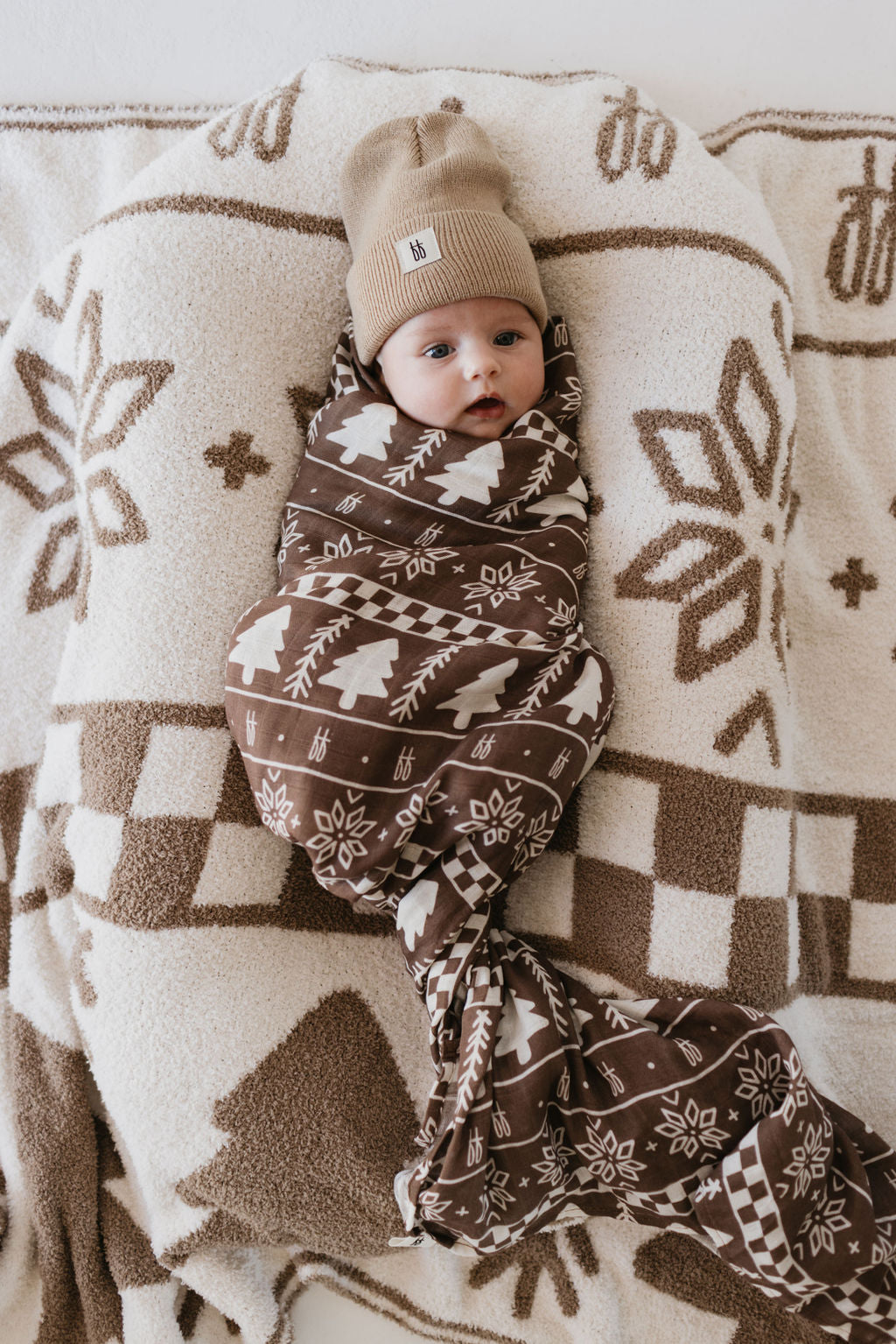 A baby wrapped in an ultra-soft Forever Fair Isle Muslin Swaddle by forever french baby, wearing a matching beige knit hat, lies on a blanket adorned with a snowflake and pine tree design. The baby's eyes are open, gazing slightly upward.