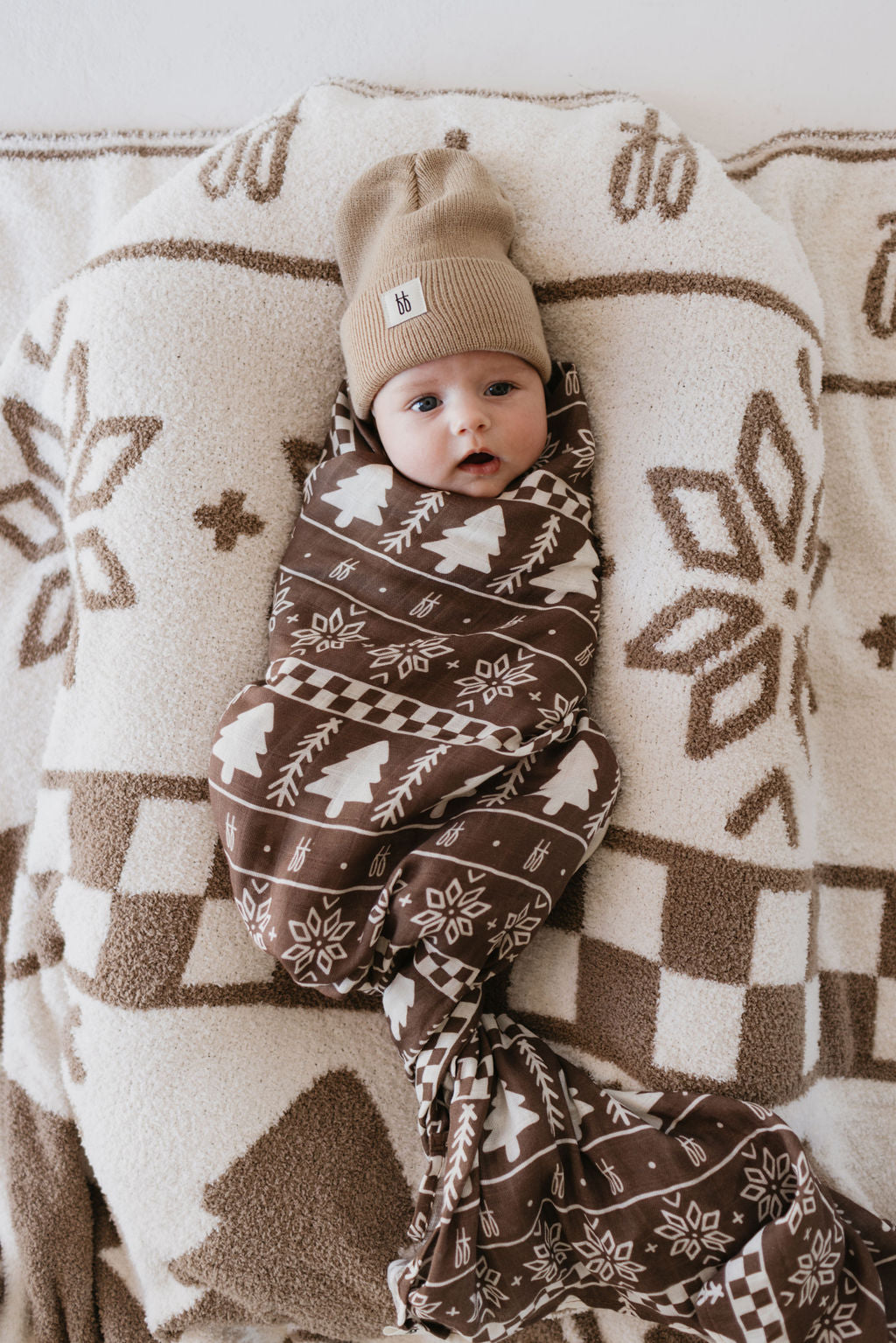 A baby wrapped in the ultra-soft "Muslin Swaddle | Forever Fair Isle" by forever french baby lies on a matching pillow. The little one, wearing a beige knit beanie and sporting a curious expression, is surrounded by geometric and floral designs on the cozy muslin swaddle and pillow.