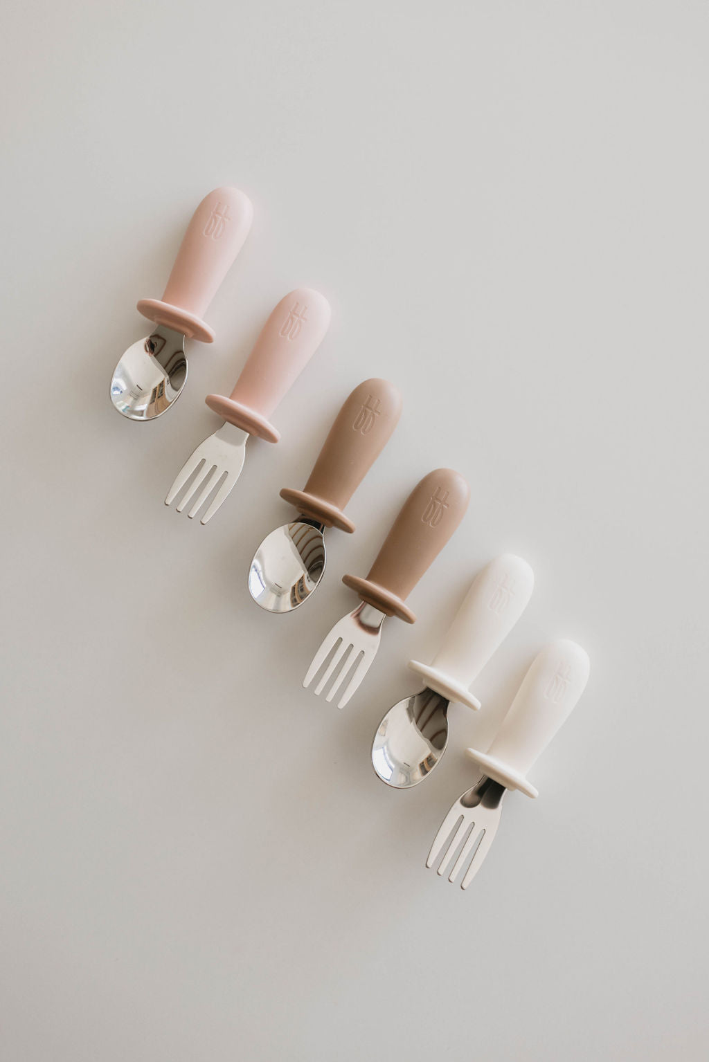 A set of Training Utensil Sets by forever french baby, featuring silicone handles in pink, beige, and white, is neatly displayed on a white surface. The set includes three forks and three spoons in alternating colors. Designed for easy gripping by small hands, these utensils encourage self-feeding for little ones.