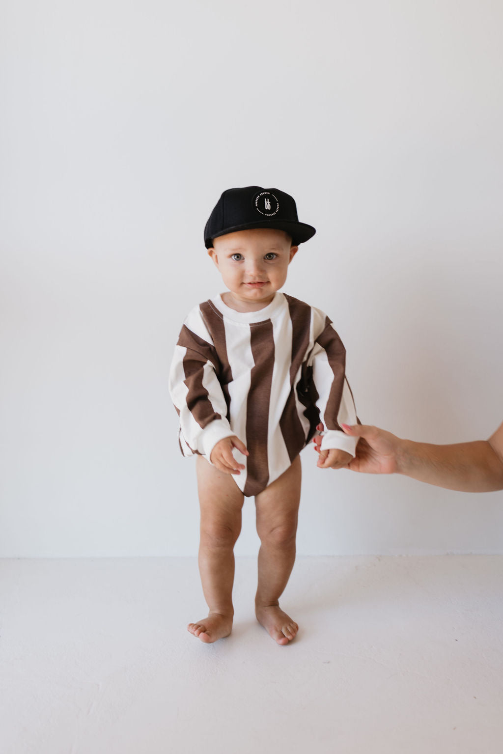 A baby wearing the "Sweatshirt Romper | Coffee Shoppe" by forever french baby is in a black cap, standing on a light-colored floor. An adult's hand gently supports the child from the side against a plain white wall backdrop.