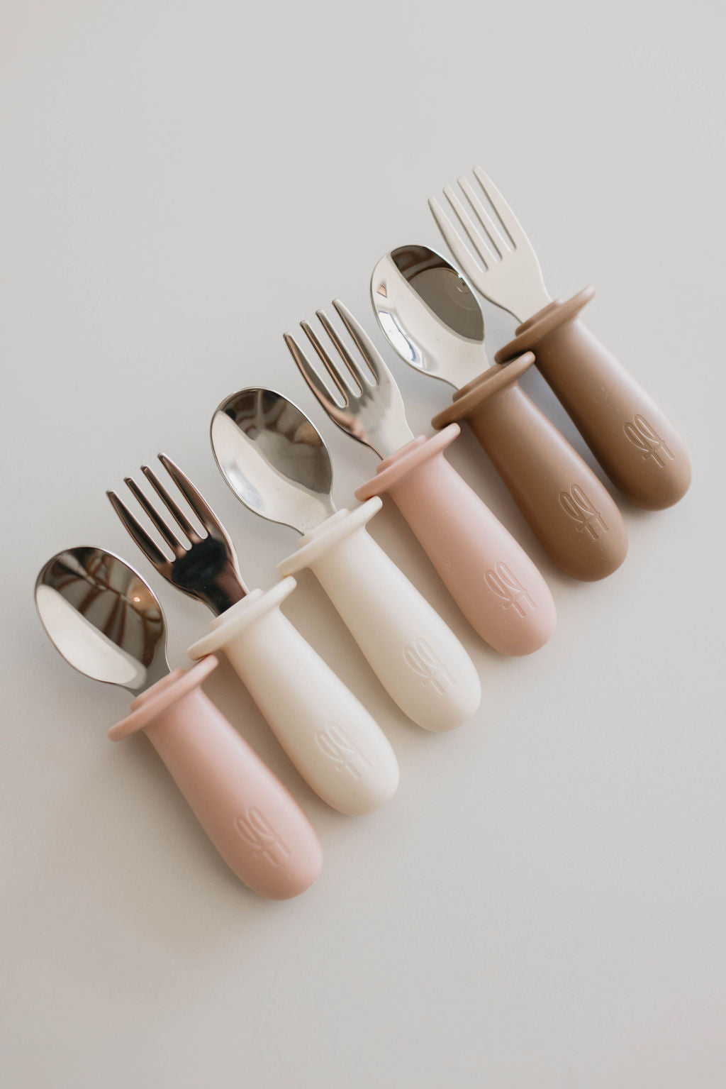 A set of forever french baby's Training Utensil Sets arranged horizontally on a white surface. The set includes three small forks and three small spoons, each with rounded silicone handles in pastel colors: pink, white, and beige. The handles feature a subtle embossed design for easy self-feed practice.