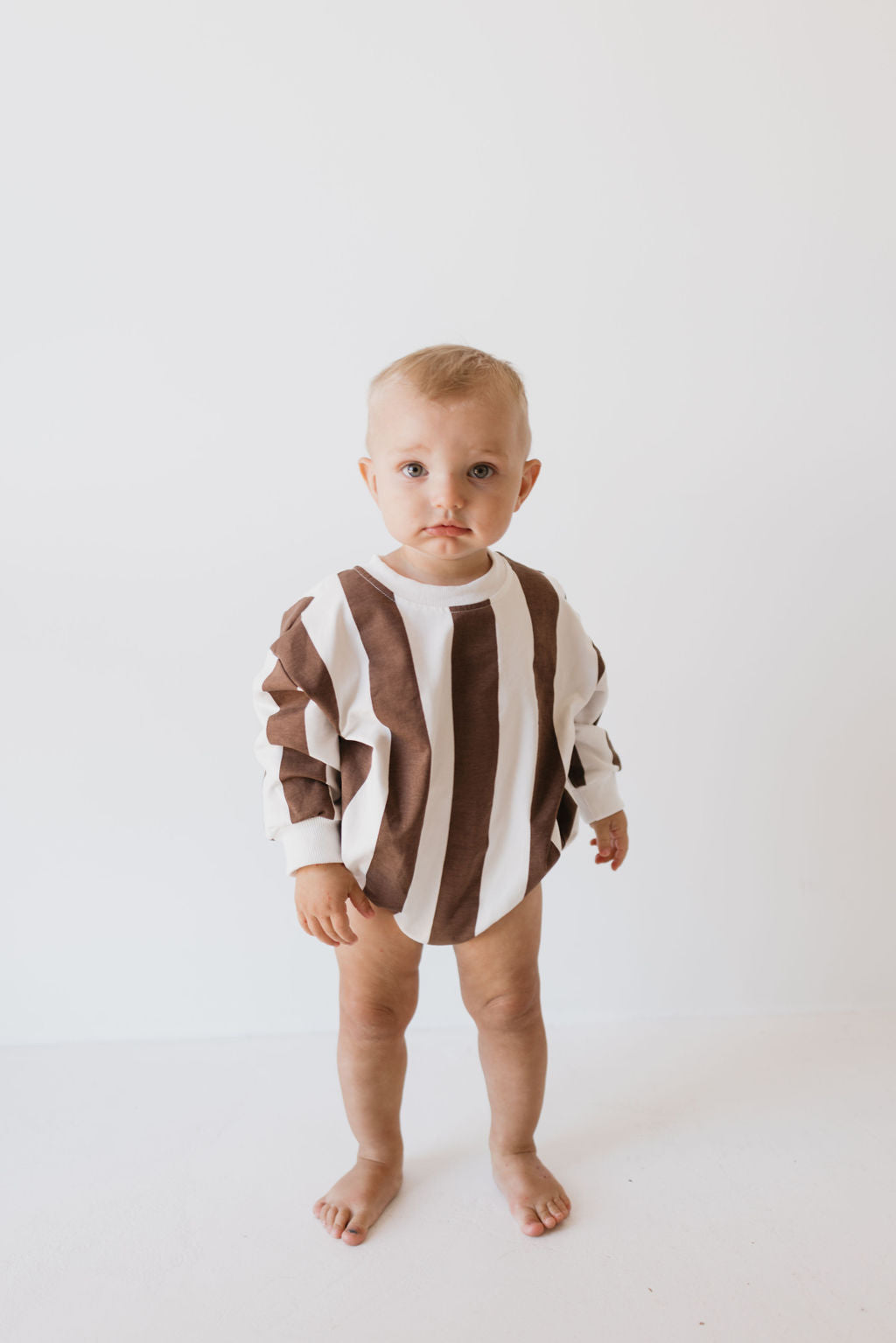 A toddler stands against a simple white backdrop wearing a brown and white striped long-sleeve shirt that evokes the cozy feel of the "Sweatshirt Romper | Coffee Shoppe" from forever french baby. The child appears with a neutral expression and is barefoot.