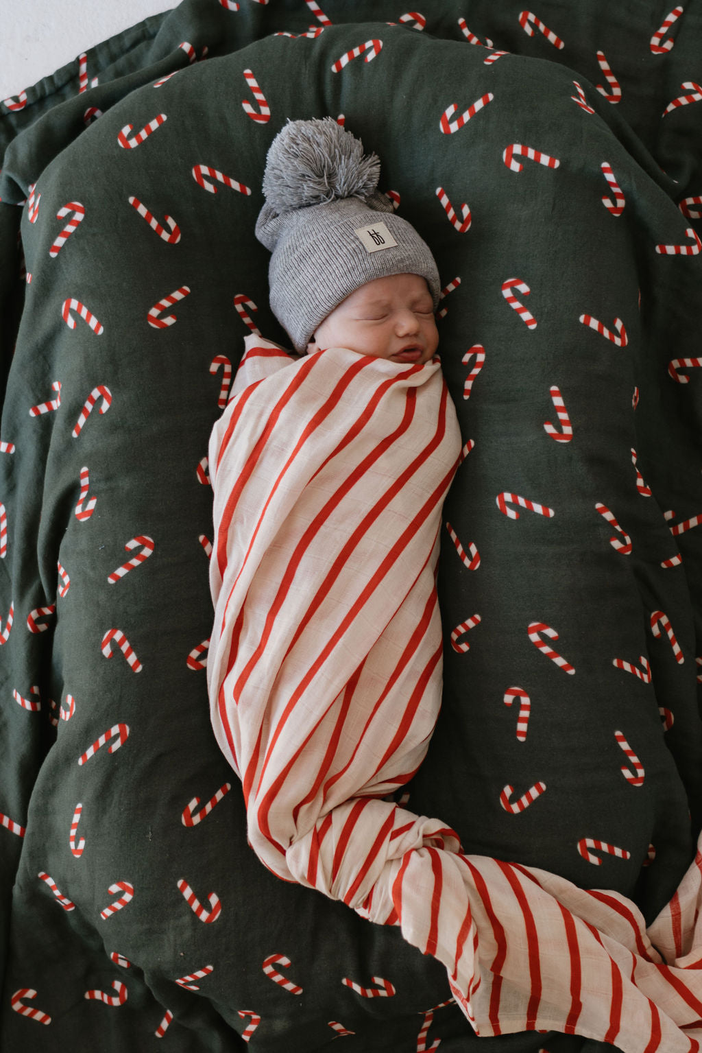 A sleeping newborn, wrapped in the Candy Cane Lane & the Claus muslin quilt by forever french baby, wears a gray knit hat with a pom-pom. The baby enjoys rest on a dark green blanket adorned with patterns that evoke the charm of Candy Cane Lane.