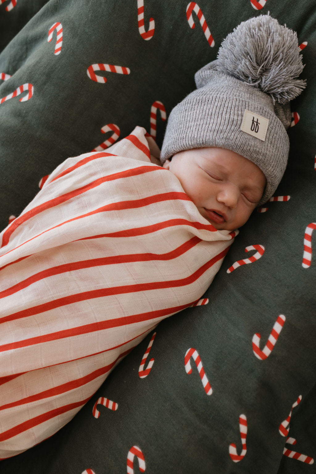 A sleeping baby is snugly wrapped in the forever french baby's Muslin Quilt | Candy Cane Lane & the Claus, perfectly nestled on a dark green pillow sprinkled with candy cane patterns—a serene scene straight out of Candy Cane Lane. The baby wears a gray knit hat topped with a playful pom-pom.