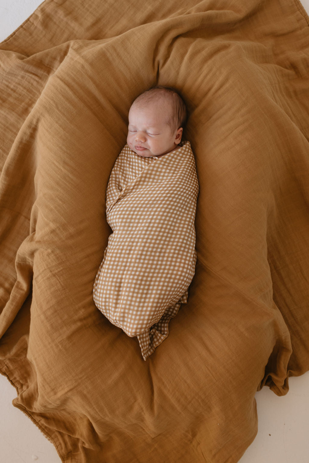 A baby peacefully sleeps swaddled in the "Muslin Swaddle | Golden Gingham" by forever french baby, nestled on a larger mustard-colored fabric. The soft, neutral tones create a warm and cozy atmosphere.