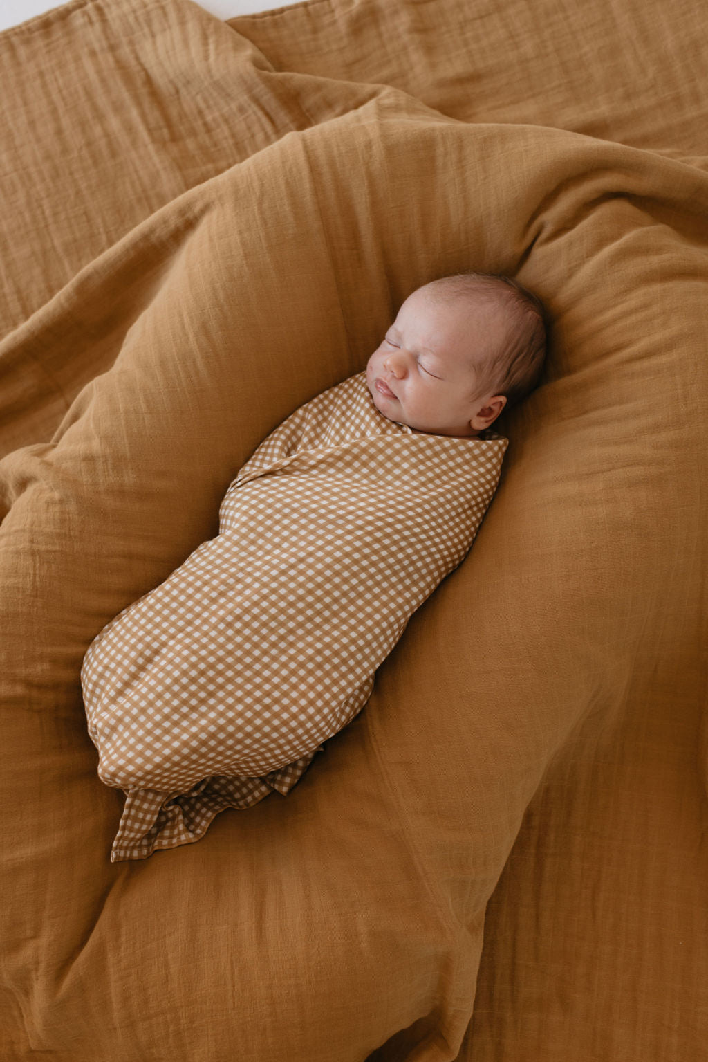 A peaceful baby sleeps swaddled in the Muslin Swaddle in Golden Gingham by forever french baby, nestled on a soft, brown cushion. The cozy scene is accentuated by the matching brown fabric, creating a serene atmosphere.