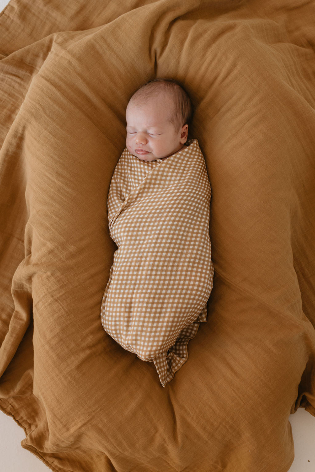 A newborn baby is peacefully sleeping, wrapped in a forever french baby Muslin Swaddle in Golden Gingham, nestled within a large, soft brown blanket. The surrounding fabric creates a cozy and warm atmosphere.