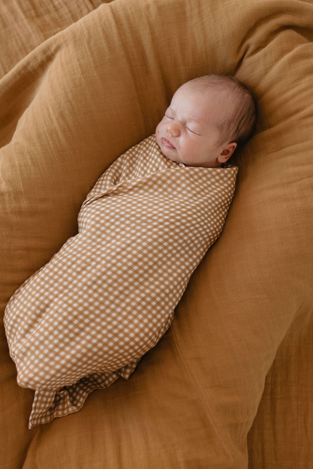 A newborn baby peacefully sleeping, wrapped in the "Muslin Swaddle | Golden Gingham" by forever french baby, resting on a soft brown cushion.