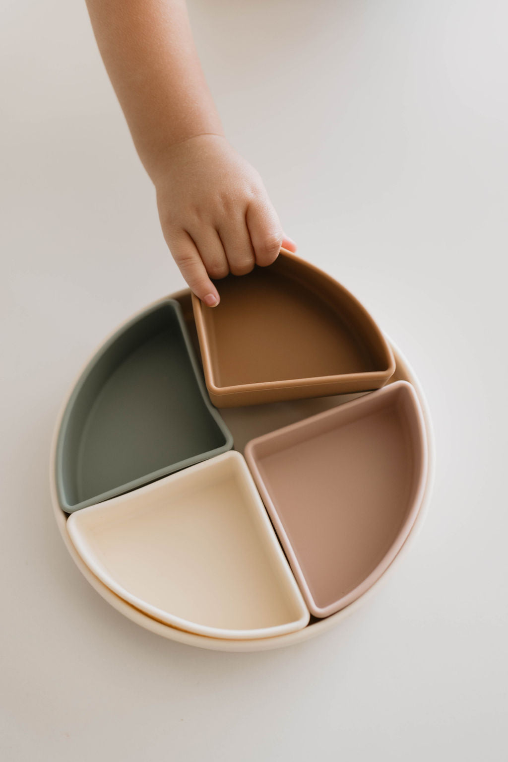 A child's hand is lifting a brown triangular section from the forever french baby Silicone Plate with Removable Dividers. The plate contains four triangular pieces in muted colors—green, cream, brown, and tan—arranged in a circular pattern on a white surface. Perfect for making mealtime fun and organized!