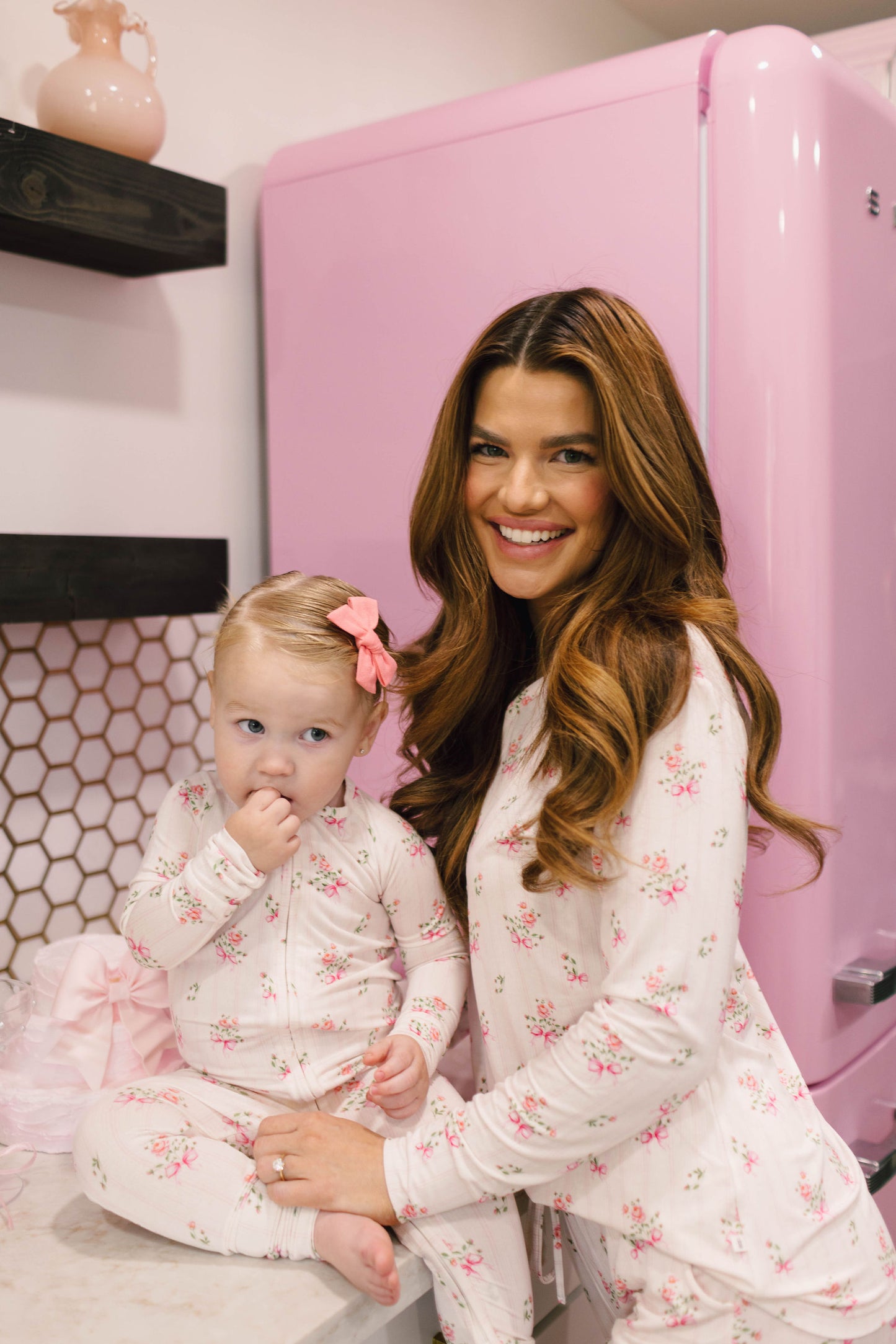 A woman with long brown hair and a child with a pink bow in her hair sit together on a white countertop. Both are wearing matching Women's Bamboo Pajamas from the Kendy Floral collection by Kendy 2. They are in a kitchen with a pink refrigerator and honeycomb-patterned backsplash. The child has her hand in her mouth.