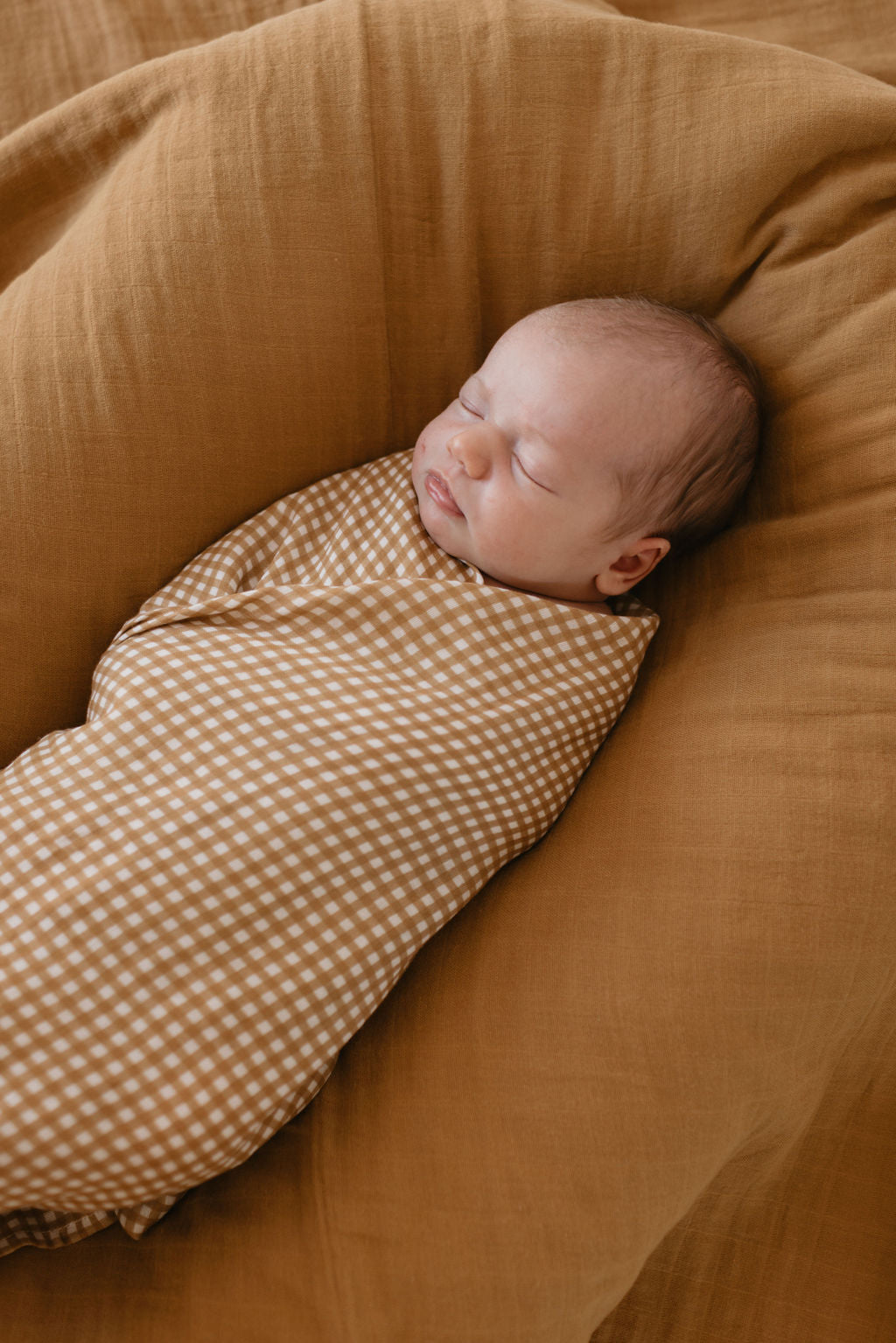 A peaceful baby sleeps wrapped in the Muslin Swaddle in Golden Gingham by forever french baby. The baby is lying on a soft mustard-colored cushion, with eyes closed and lips slightly parted, exuding warmth and serenity.