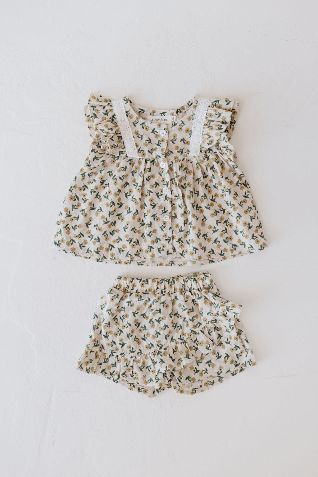 A delightful toddler outfit by forever french baby, the Two Piece Skort Set in Tulip Fields, is crafted from 100% cotton. It features a sleeveless blouse with frilled shoulders and matching shorts. The fabric boasts small green leaves and yellow tulips against a light beige background, all beautifully arranged on a white surface.