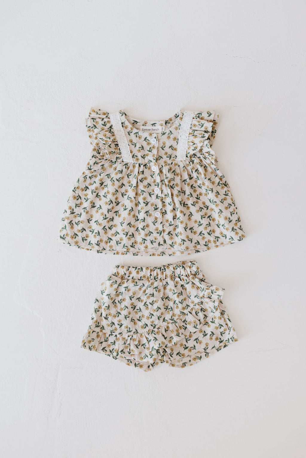 The "Tulip Fields" Two Piece Skort Set from forever french baby is laid out on a white background. It features a sleeveless top with ruffled sleeves and lace detailing, paired with shorts. The fabric is 100% cotton and showcases a delicate floral pattern in muted tones.