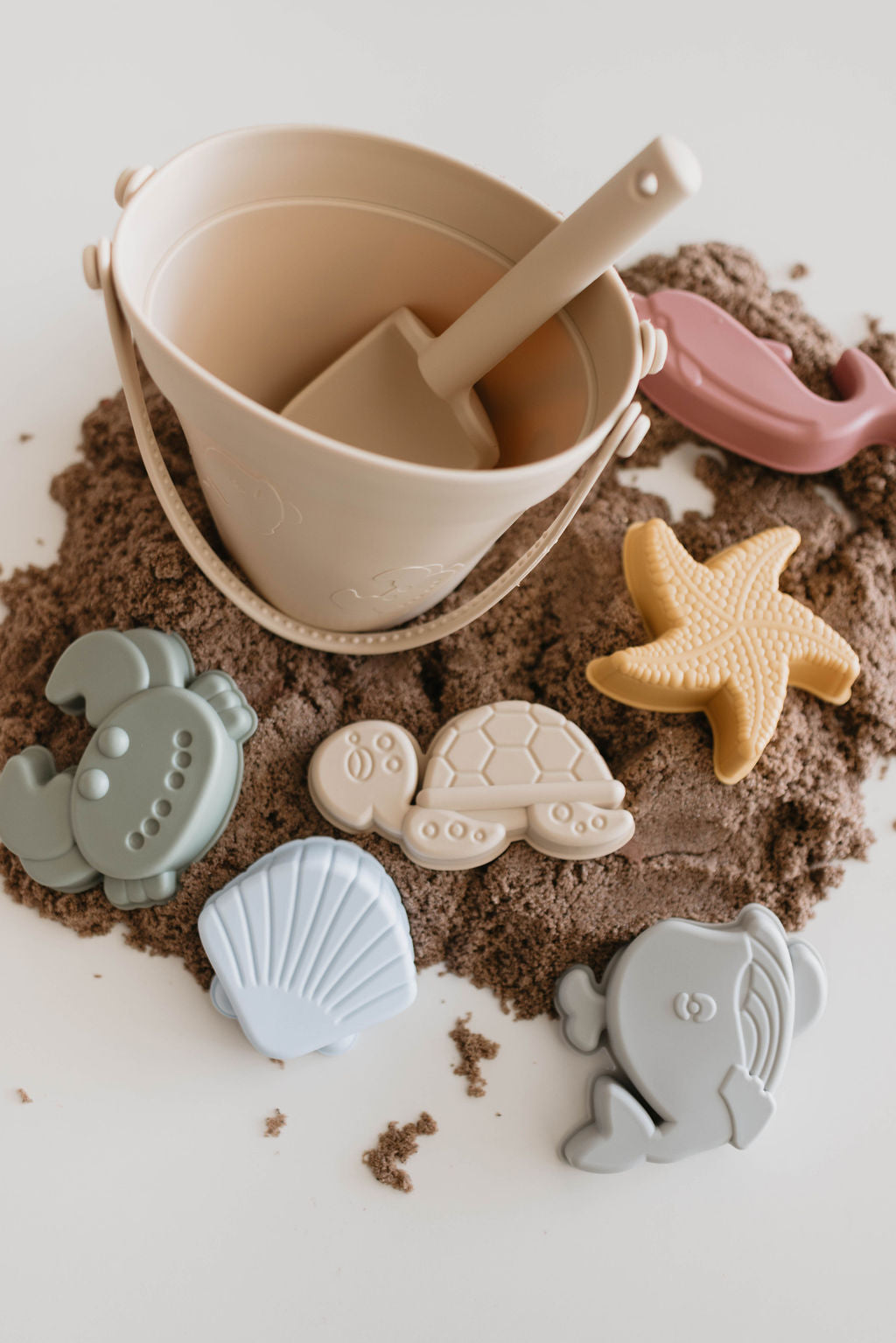 A beige Silicone Sand Bucket from forever french baby, complete with a matching shovel, rests on a mound of sand. Surrounding it are ocean creature sand molds in the shapes of a turtle, starfish, crab, fish, and clam shell, along with an additional pink toy shovel — the perfect beach companion from the 8 Piece set.