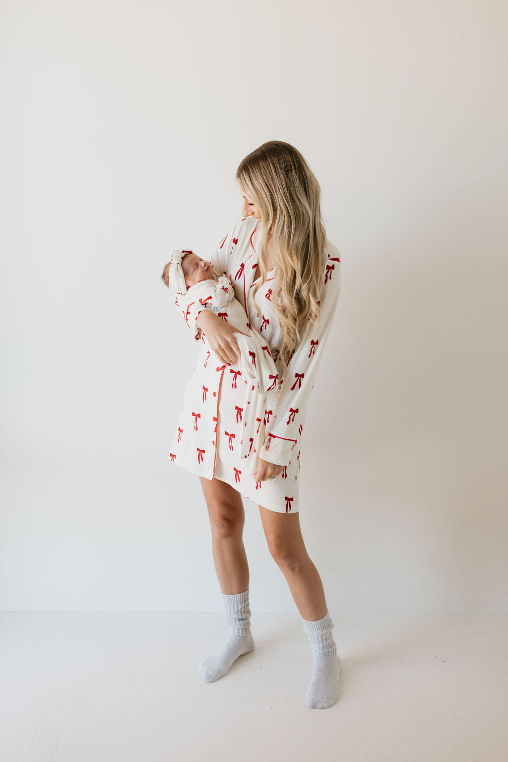 A woman with long blonde hair stands against a white background, wearing the Bamboo Knotted Gown in the Ribbons & Bows pattern from forever french baby, paired with gray socks. She cradles her newborn wrapped in a matching hypo-allergenic gown. Holding the baby close, she looks down lovingly.