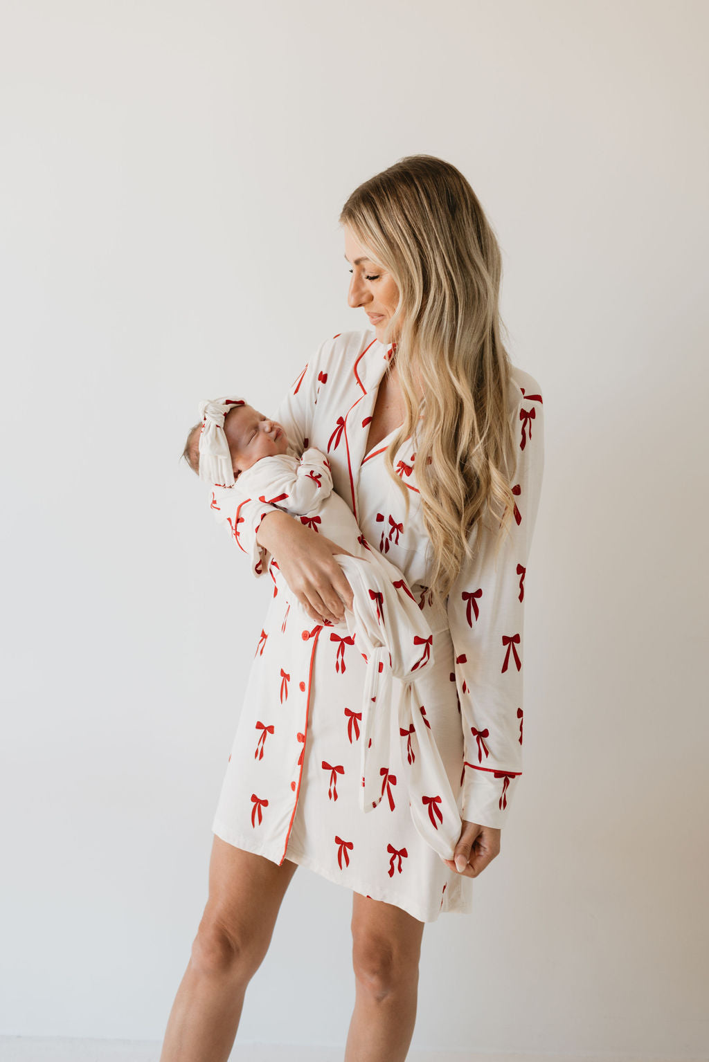 A woman with long blonde hair, dressed in the Bamboo Knotted Gown featuring the charming Ribbons & Bows design from forever french baby, lovingly holds a newborn wrapped in hypo-allergenic bamboo clothing. They stand against a plain light-colored background, embodying both comfort and newborn essentials.