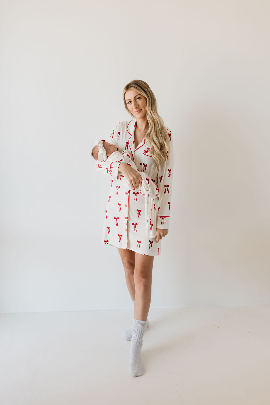 A woman with long blonde hair stands barefoot on a light background, wearing the "Women's Bamboo Sleeping Dress | Ribbons & Bows" by forever french baby. She is holding a baby wrapped in a hypo-allergenic white blanket, and her expression is relaxed and gentle.