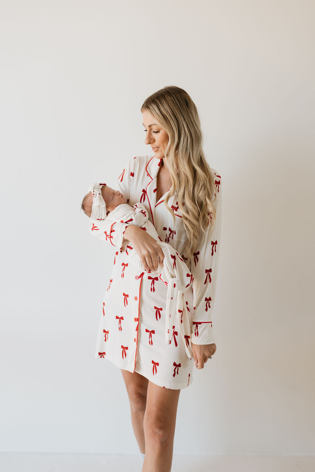 A woman wearing a Women's Bamboo Sleeping Dress from forever french baby's 'Ribbons & Bows' collection, which features a white design with red bows reminiscent of a bamboo night gown, cradles a baby wrapped in a matching blanket. They pose against a simple white background. The woman has long blonde hair and smiles softly at the baby.