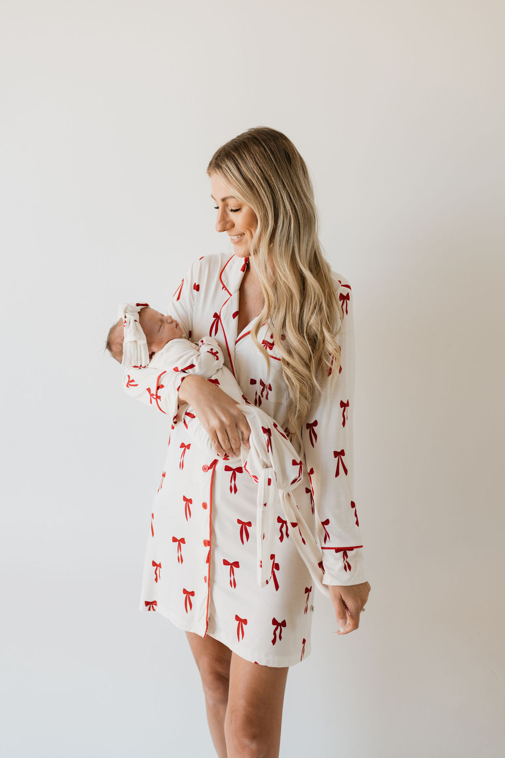 A woman with long blonde hair holds a newborn baby, both wearing matching outfits made by forever french baby. The clothes are crafted from hypoallergenic fabric and feature the Ribbons & Bows pattern. The woman beams as she looks lovingly at the baby in her arms, standing against a simple light background.