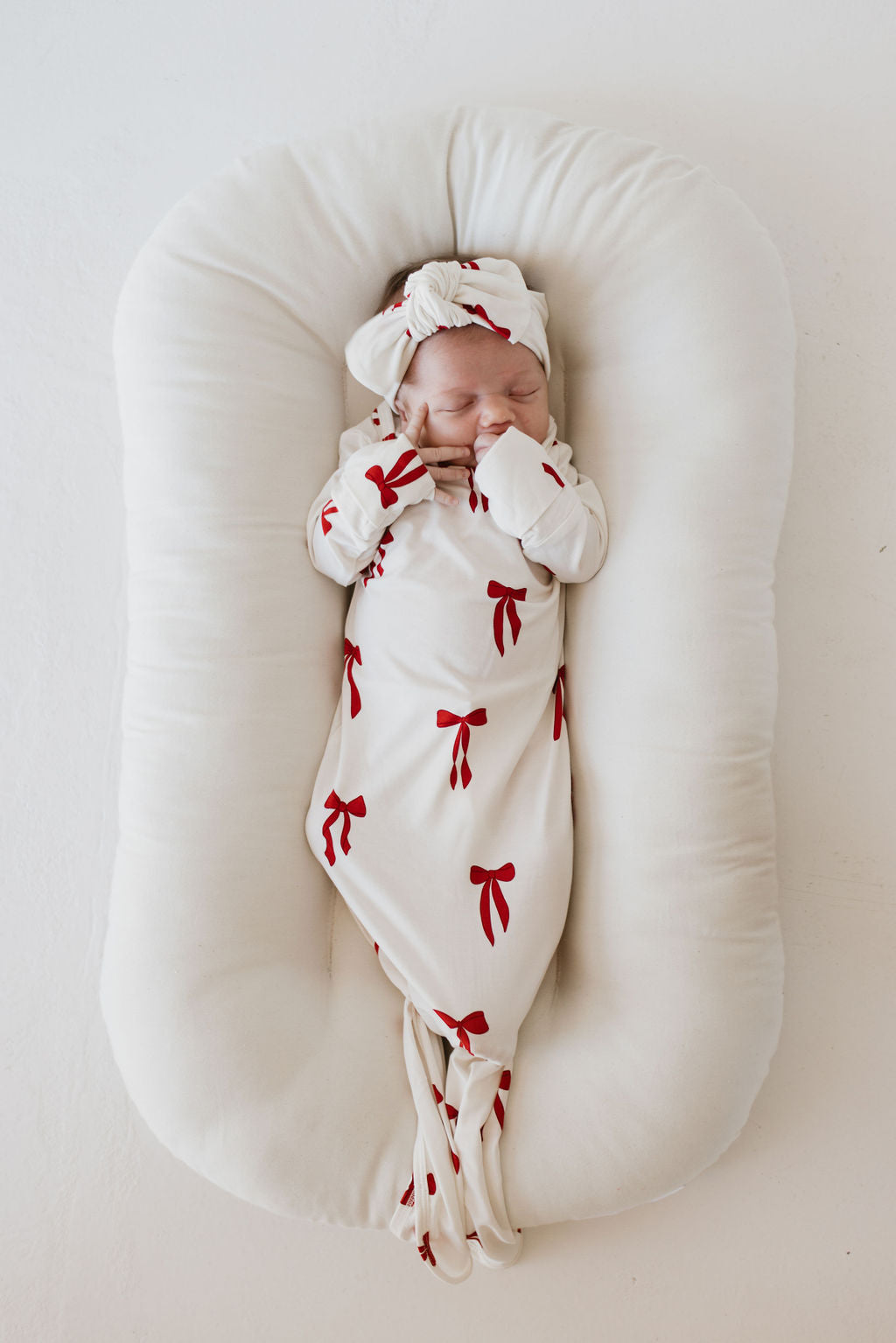 A sleeping baby dressed in forever french baby's Bamboo Head Wrap | Ribbons & Bows, featuring a charming white outfit adorned with cute red bows, is lying peacefully on a soft, white baby lounger, with their hands resting near their face.