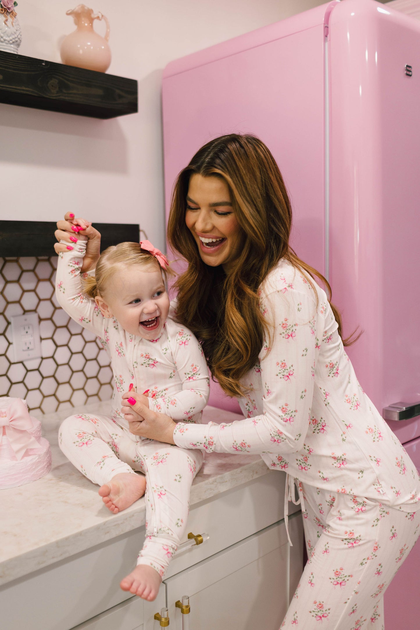 A woman and a young girl, both dressed in matching Women's Bamboo Pajamas from the Kendy Floral collection by Kendy 2, are happily playing in a kitchen. The woman has long brown hair and is smiling warmly at the girl, who is laughing with her hand raised. The kitchen features a pink refrigerator and decorative shelves.