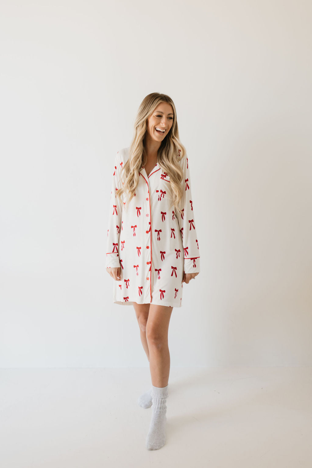 A woman with long blonde hair is wearing the Women's Bamboo Sleeping Dress in the Ribbons & Bows pattern from forever french baby. She's standing in a bright, minimalistic space, smiling with her hands by her sides and wearing gray socks. Her attire exudes comfort and sustainability, capturing the charm of this eco-friendly bamboo night gown.