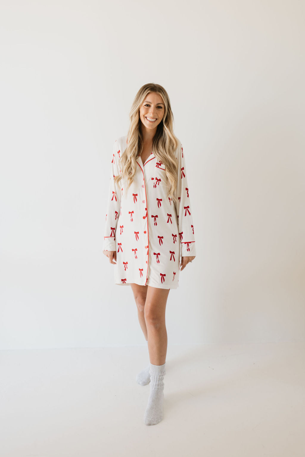 A person with long hair stands against a plain background, wearing the eco-friendly Women's Bamboo Sleeping Dress in the Ribbons & Bows design from forever french baby. They are smiling and looking at the camera.