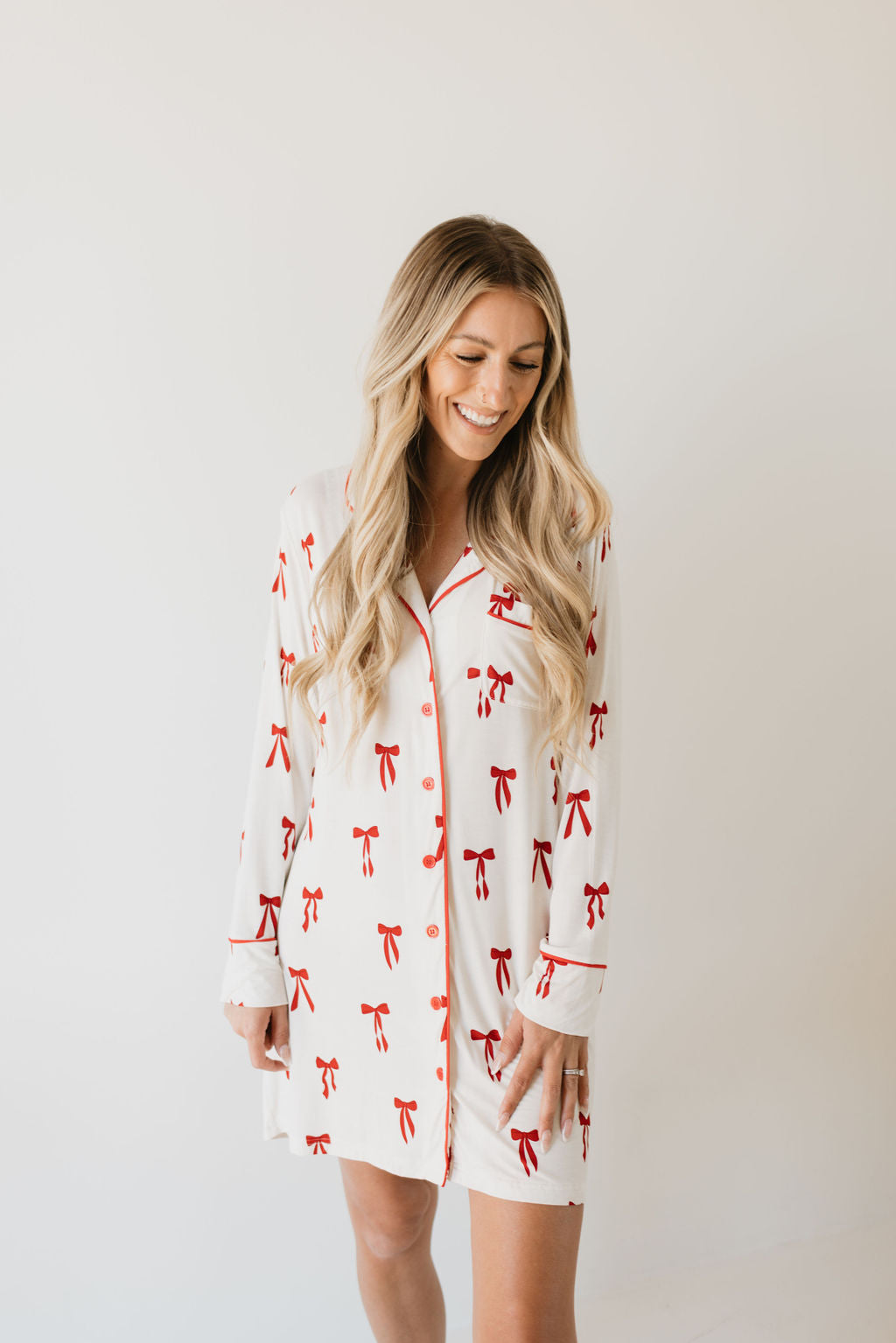 A smiling woman with long blonde hair models the Women's Bamboo Sleeping Dress from forever french baby, featuring an eco-friendly white design adorned with red ribbons and matching bows, against a simple light backdrop.