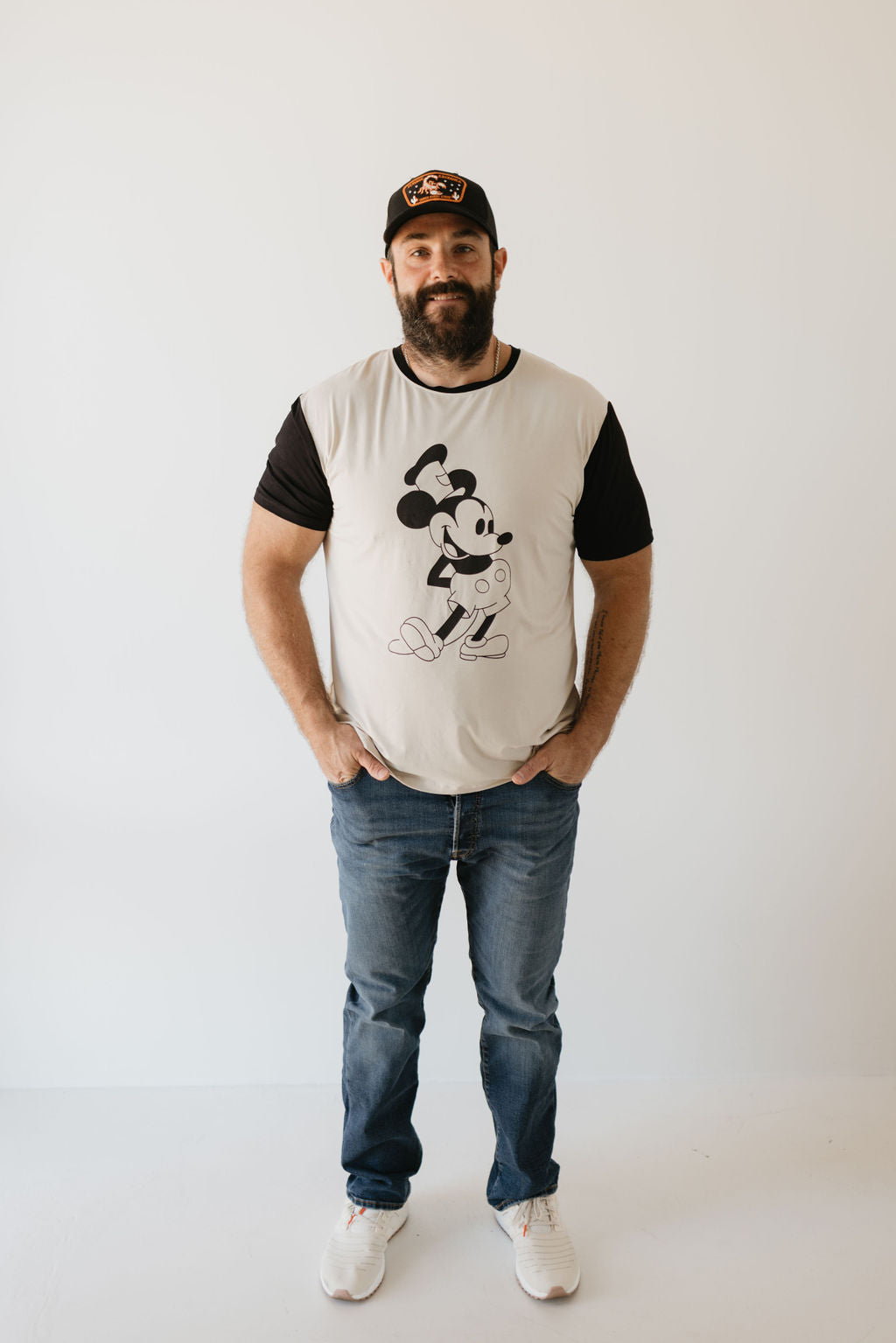 A man stands against a white background, wearing an Adult Bamboo Tee featuring Steamboat Willie by forever french baby, jeans, white sneakers, and a cap.