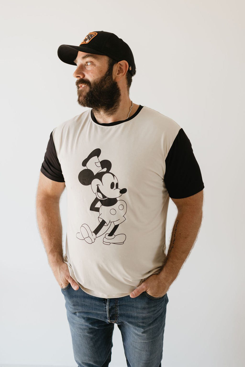 A bearded man wearing a baseball cap and an Adult Bamboo Tee from forever french baby, which features a vintage Steamboat Willie print, stands against a plain background. With his hands in his pockets, he casually gazes off to the side.