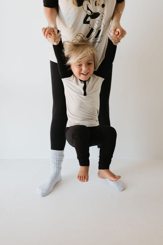 A young child with blonde hair, dressed in breathable attire, joyfully swings while holding the hands of an adult behind them. The adult, in black leggings and gray socks, wears a forever french baby shirt featuring a Steamboat Willie illustration from their Bamboo Two Piece Pajamas collection, perfectly complementing the playful moment.