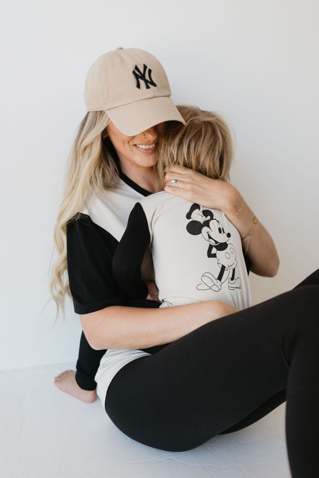 A woman in a beige cap and black leggings holds a young child dressed in forever french baby's Bamboo Two Piece Pajamas featuring the Steamboat Willie design. They are sitting against a white wall, with the child nestled comfortably in the woman's arms.