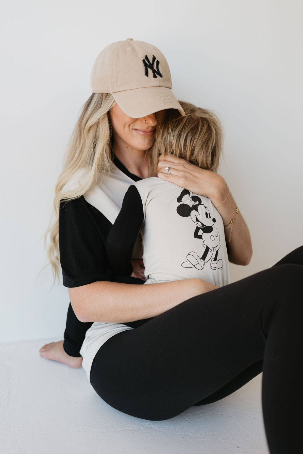 A woman in a baseball cap sits on the floor, lovingly hugging a child who is wearing the Bamboo Two Piece Pajamas | Steamboat Willie by forever french baby. The woman has long blonde hair and wears hypo-allergenic clothing—a black and white shirt with black leggings. Their expressions are soft and tender.